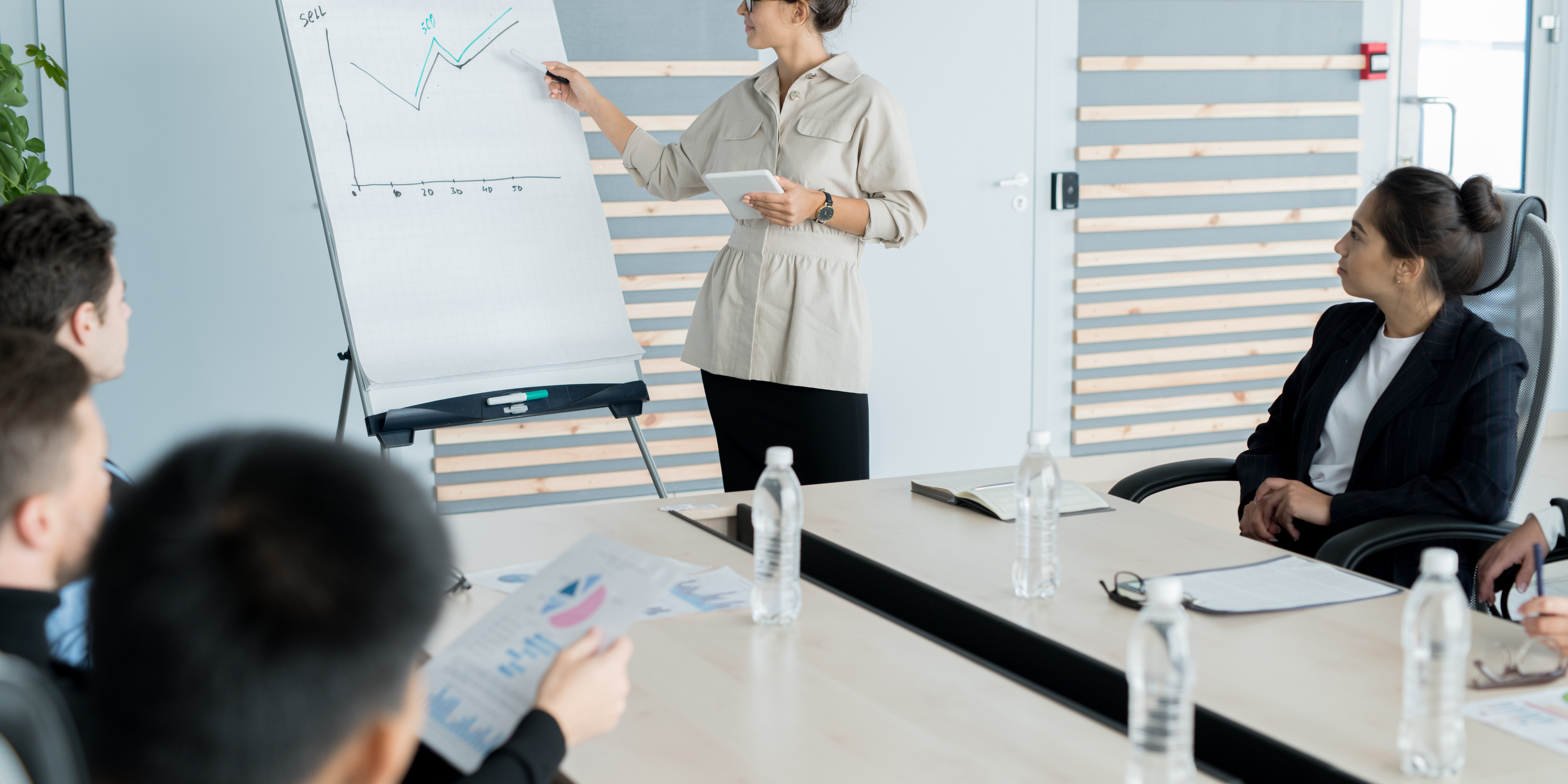 Young sales coach with hair bun wearing glasses holding presentation and explaining graph to business people at training class